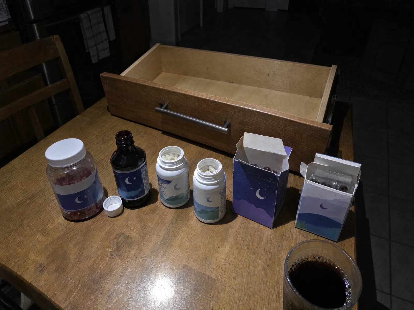 A kitchen drawer pulled out at 3am with sleep aid bottles lined up on the table and a cold cup of coffee